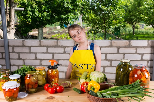 Ragazza giovane indossando il grembiule in piedi fuori vicino al giardino di un muro di mattoni e preparare verdure fresche per la conservazione o il decapaggio in barattoli di vetro