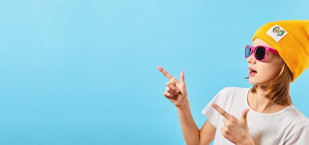 Ragazza giovane isolata sopra la parete blu. Adolescente, mostrando copyspace indicando. Hipster Teen in occhiali alla moda che indossa un cappello lavorato a maglia. ampia foto