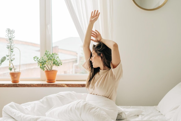 Ragazza giovane si allunga la mattina dopo essersi svegliata mentre è seduta sul letto in una camera da letto accogliente