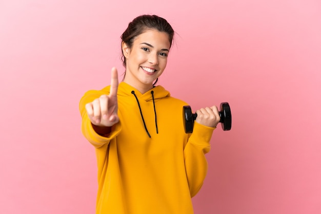 Ragazza giovane sport facendo sollevamento pesi su sfondo rosa isolato che mostra e alzando un dito