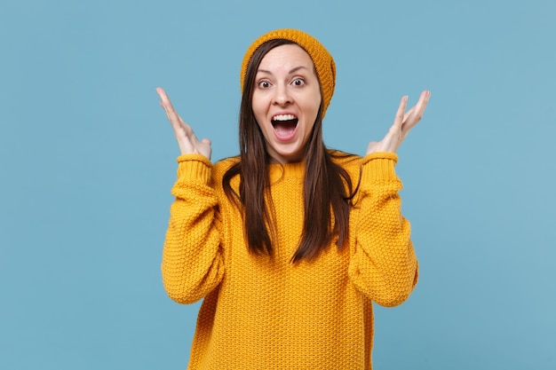 Ragazza giovane stupita in maglione giallo e cappello in posa isolata su sfondo blu ritratto in studio. Persone sincere emozioni concetto di stile di vita. Mock up spazio di copia. Tenere la bocca aperta allargando le mani.