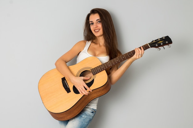 Ragazza glamour con la chitarra in mano che guarda la telecamera e sorride in studio su sfondo grigio