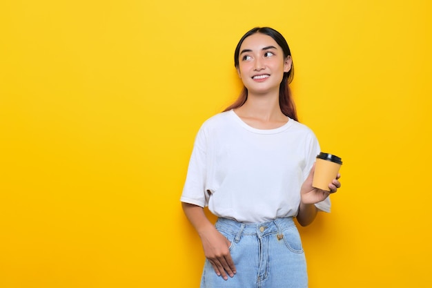 Ragazza graziosa allegra in maglietta bianca che tiene una tazza di caffè e distoglie lo sguardo nello spazio vuoto isolato su sfondo giallo