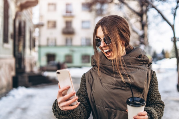 Ragazza graziosa che cammina sulla strada con il telefono e la bevanda calda.