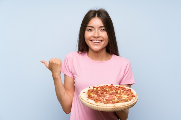 Ragazza graziosa che tiene una pizza sopra la parete blu isolata che indica il lato per presentare un prodotto