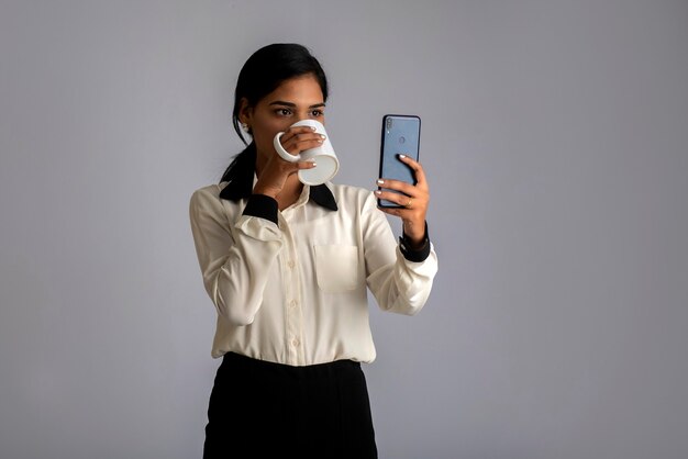 Ragazza graziosa con una tazza di tè o caffè utilizza lo smartphone in posa sul grigio.