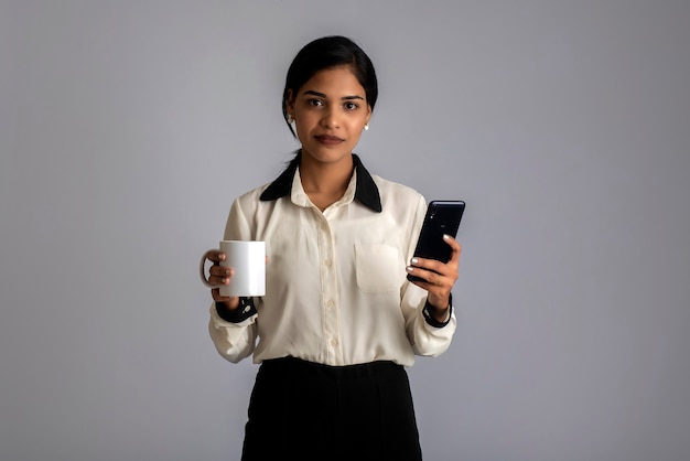 Ragazza graziosa con una tazza di tè o caffè utilizza lo smartphone in posa sul grigio.