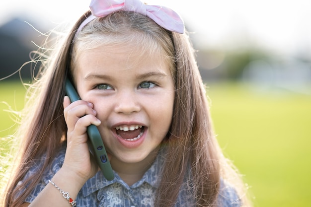 Ragazza graziosa del bambino che parla sul telefono cellulare che sorride felicemente all'aperto in estate.