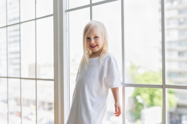 Ragazza graziosa felice che porta vestito bianco in bianco vicino alla grande finestra