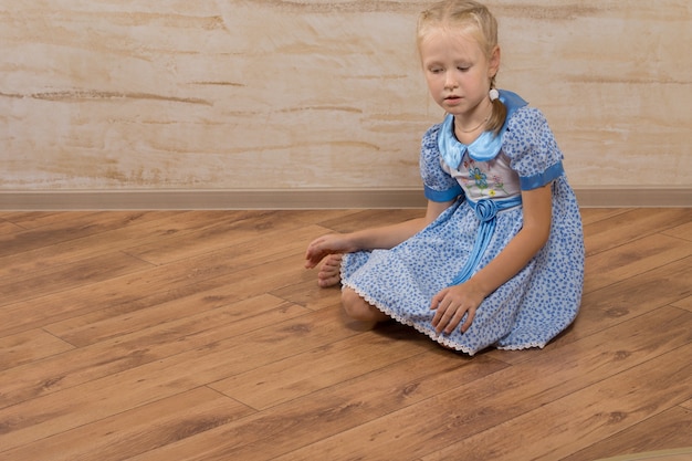 Ragazza graziosa seria sul vestito blu che si siede sul pavimento di legno di Brown.