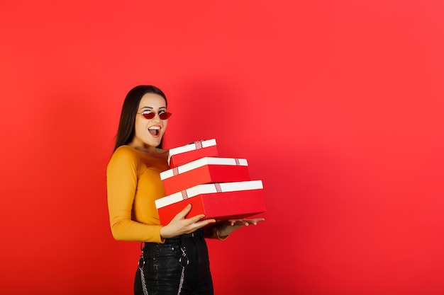 Ragazza graziosa sorridente emotiva che tiene pila di scatole regalo isolato sulla superficie rossa con spazio di copia.