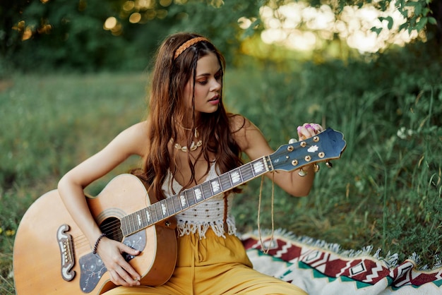 Ragazza hippie donna che suona la chitarra in abiti ecofriendly seduti per terra fuori nella natura in autunno guardando il tramonto