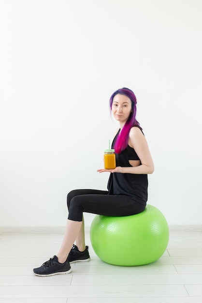 Ragazza hipster di razza mista con i capelli colorati seduto su un fitball verde e con in mano un frullato proteico di banana su uno sfondo bianco. Mangiare sano e concetto di esercizio.