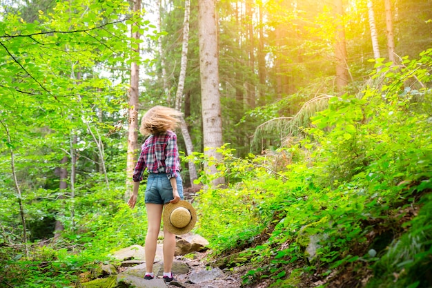 Ragazza hipster in montagna. Donna alla moda in cappello di paglia e camicia a scacchi nella foresta. Concetto di voglia di viaggiare. Viaggiare in estate. legno misterioso. Natura meravigliosa.