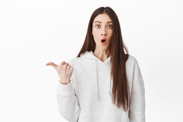 Ragazza impressionata e sorpresa che guarda con soggezione, indicando da parte il logo a sinistra, mostrando un nuovo fantastico sconto promozionale, dimostrando l'offerta del negozio, in piedi sul muro bianco