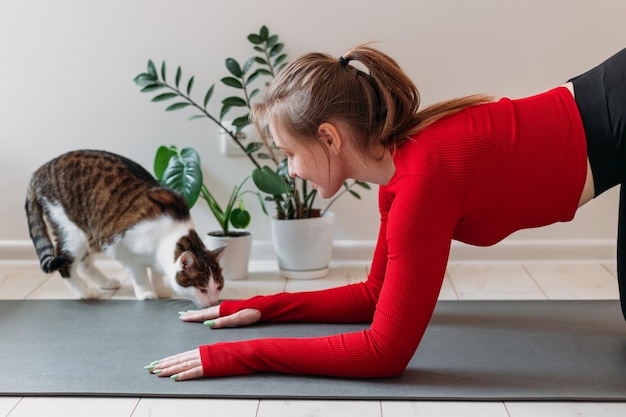 Ragazza in abbigliamento sportivo rosso con simpatico gatto divertente che pratica yoga a casa Allenamento indoor e outdoor Concetto di sport e stile di vita sano e attivo