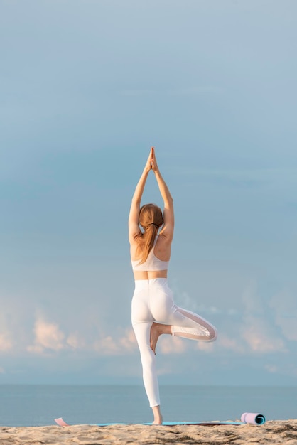 Ragazza in abiti sportivi bianchi che praticano yoga posa dell'albero sulla spiaggia Vrikshasana Giovane donna che fa yoga con il mare dietro