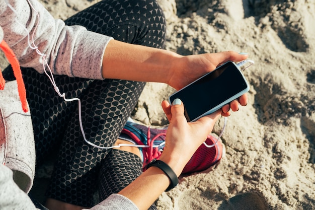 Ragazza in abiti sportivi che ascolta la musica in cuffia su un telefono cellulare sulla spiaggia al mattino