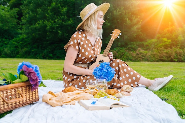 Ragazza in abito a pois e cappello seduto su una coperta da picnic in maglia bianca suona l'ukulele e beve vino