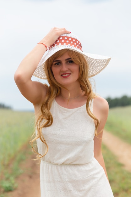 Ragazza in abito bianco e cappello su uno sfondo di campo. La ragazza con un cappello si alza, si rilassa all'aperto, si diverte, concetto di armonia.