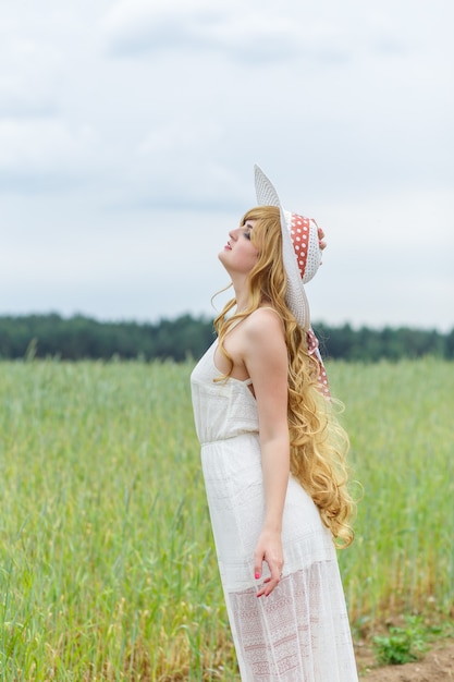 Ragazza in abito bianco e cappello su uno sfondo di campo. La ragazza con un cappello si alza, si rilassa all'aperto, si diverte, concetto di armonia.