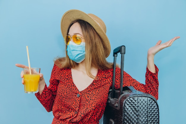 Ragazza in abito, cappello e occhiali da sole con mascherina medica sul viso e cocktail nelle sue mani.