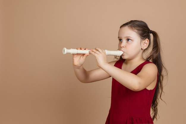 Ragazza in abito rosso che guarda lontano suona la melodia sul flauto che soffia aria nel condotto sfondo beige Impara a suonare uno strumento musicale a fiato