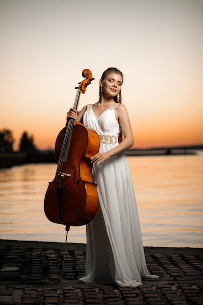 Ragazza in bel vestito bianco con un violoncello si trova in riva al lago al tramonto