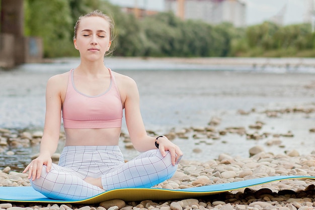Ragazza in buona salute che si rilassa mentre meditando e facendo esercizio di yoga nella bella natura sulla riva del fiume