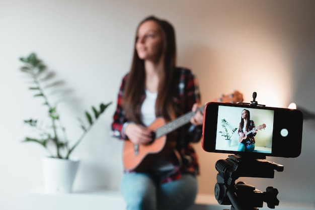 Ragazza in camicia e jeans scrive un vlog al telefono sul gioco delle ukulele.