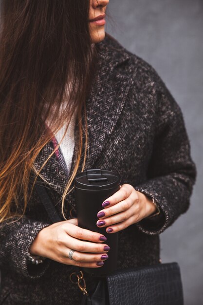 Ragazza in cappotto elegante con bella mano di manicure che tiene un bicchiere vuoto nero su un muro grigio