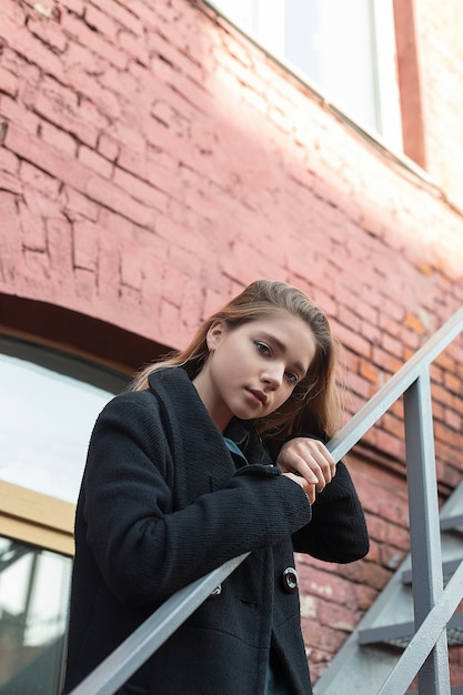 Ragazza in cappotto nero che sta sulle scale con il muro di mattoni su fondo. solitudine.