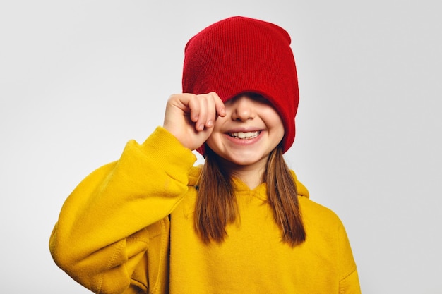 Ragazza in felpa con cappuccio gialla che copre gli occhi con un cappello rosso che sorride alla telecamera su bianco
