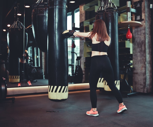 Ragazza in forma in abiti sportivi e treni guantoni da boxe contro un sacco da boxe in una palestra buia