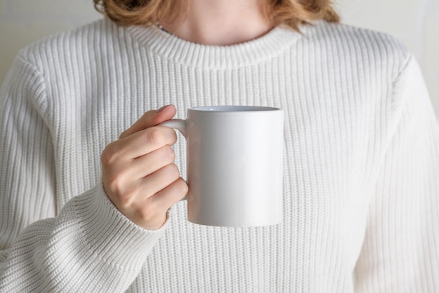 Ragazza in maglione bianco che tiene la tazza da caffè in porcellana bianca finta da vicino