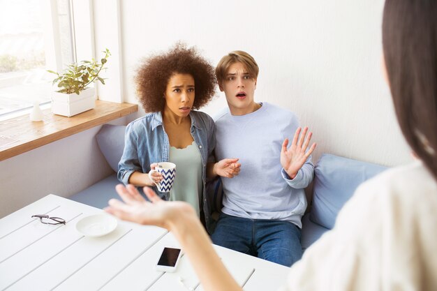 Ragazza in maglione bianco è in piedi davanti ai suoi amici e agitando con la mano. La ragazza afroamericana e un ragazzo stanno itting sul sofà. Sembrano sabbia confusa infelice.