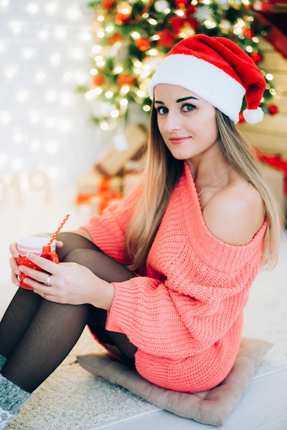 Ragazza in maglione e calze seduta sullo sfondo di un albero di Natale con una tazza in mano e un tubo da cocktail