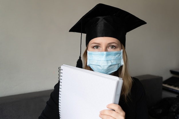 Ragazza in maschera medica con cappello da abito da laurea che tiene in mano un taccuino su sfondo grigio