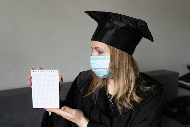 Ragazza in maschera medica con cappello da abito da laurea che tiene un piccolo taccuino su sfondo grigio