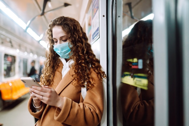 Ragazza in mascherina medica sterile protettiva sul viso con un telefono in un vagone della metropolitana. Donna che usa il telefono per cercare notizie sul coronavirus. Il concetto di prevenire la diffusione dell'epidemia.