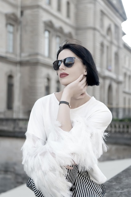 Ragazza in occhiali da sole posa sulla facciata della casa a parigi francia Donna con labbra rosse e capelli castani Modello in abiti alla moda su strada Accessorio di moda visage concept Stile look di bellezza