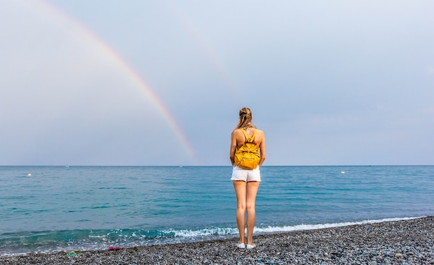 Ragazza in pantaloncini e con uno zaino giallo sulla spiaggia a guardare un arcobaleno