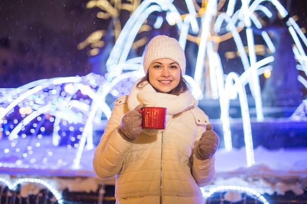 Ragazza in piedi sopra inverno natale città sfondo neve cumuli di neve, sta in piedi con il cappello caldo della giacca, tiene una tazza di tè con bevande calde.