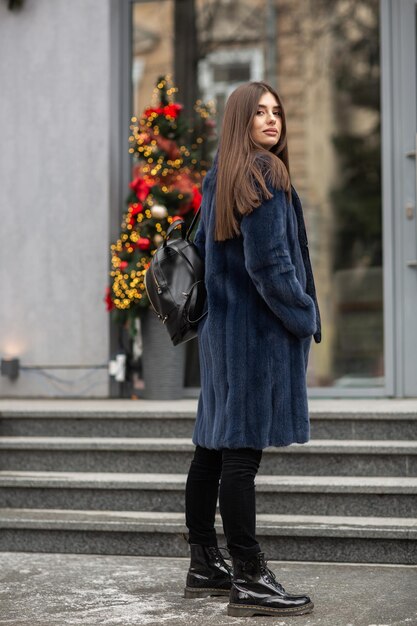 Ragazza in posa su strada su sfondo invernale Giovane donna affascinante e divertente con un sorriso che indossa un cappotto di pelliccia lungo grigio elegante Pelliccia e concetto di moda Bella gente