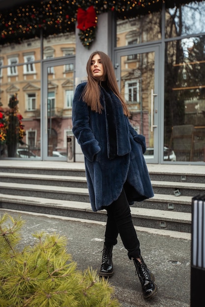 Ragazza in posa su strada su sfondo invernale Giovane donna affascinante e divertente con un sorriso che indossa un elegante cappotto di pelliccia lungo blu Pelliccia e concetto di moda