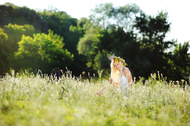 Ragazza in prendisole bianche e una corona di fiori sulla sua testa che si siede nell'erba