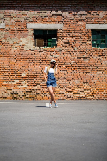 Ragazza in tuta di jeans, una maglietta bianca e un cappello leggero sullo sfondo di un edificio in mattoni sotto il sole splendente