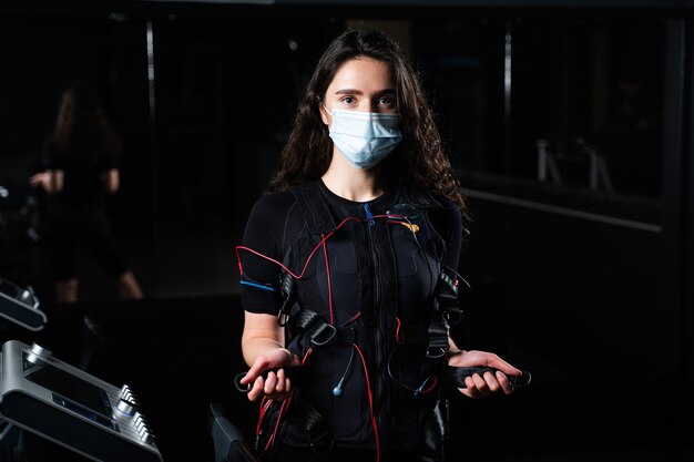 Ragazza in tuta EMS e mascherina medica in palestra. Protezione dal coronavirus covid-19. Allenamento sportivo in tuta di stimolazione muscolare elettrica.