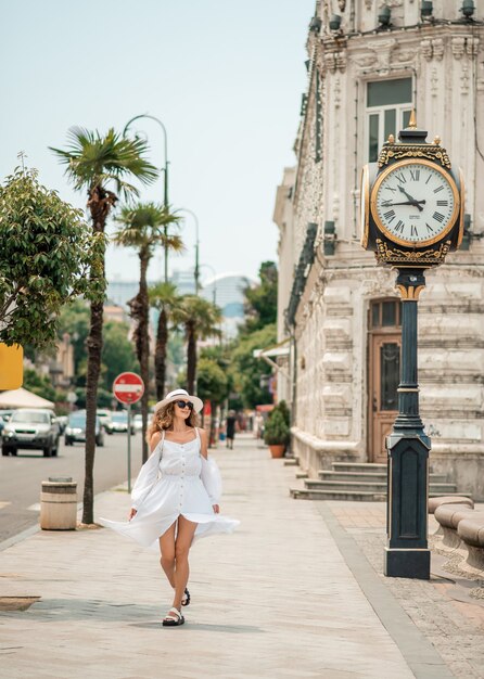 Ragazza in un abito lungo bianco, camminando vicino all'orologio. Modello di moda abbastanza sexy nel paesaggio urbano