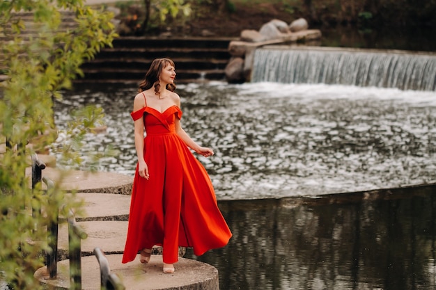 Ragazza in un lungo abito rosso vicino al lago nel parco al tramonto.